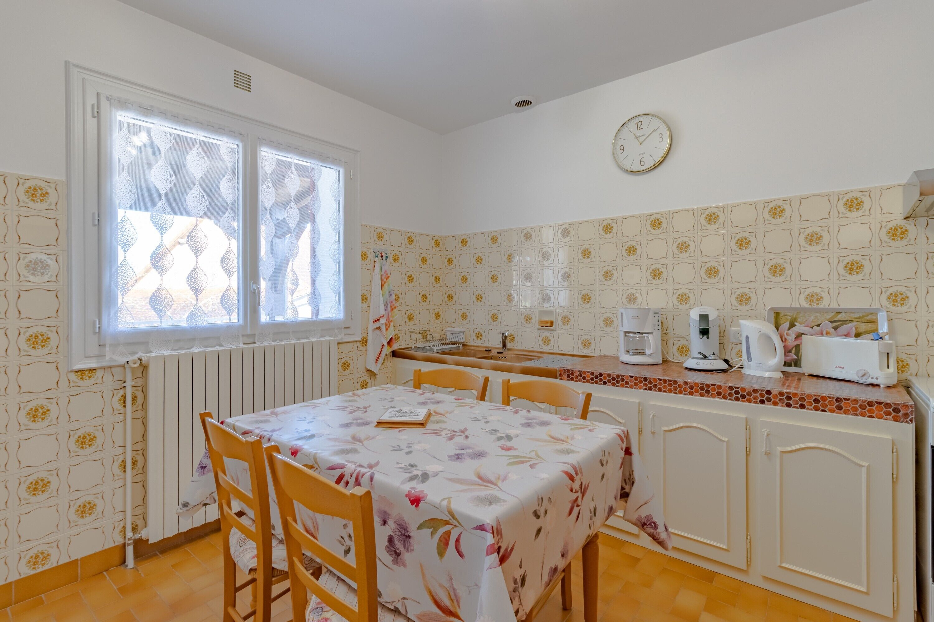 A kitchen with a dining table, chairs, a clock, and a coffee maker.