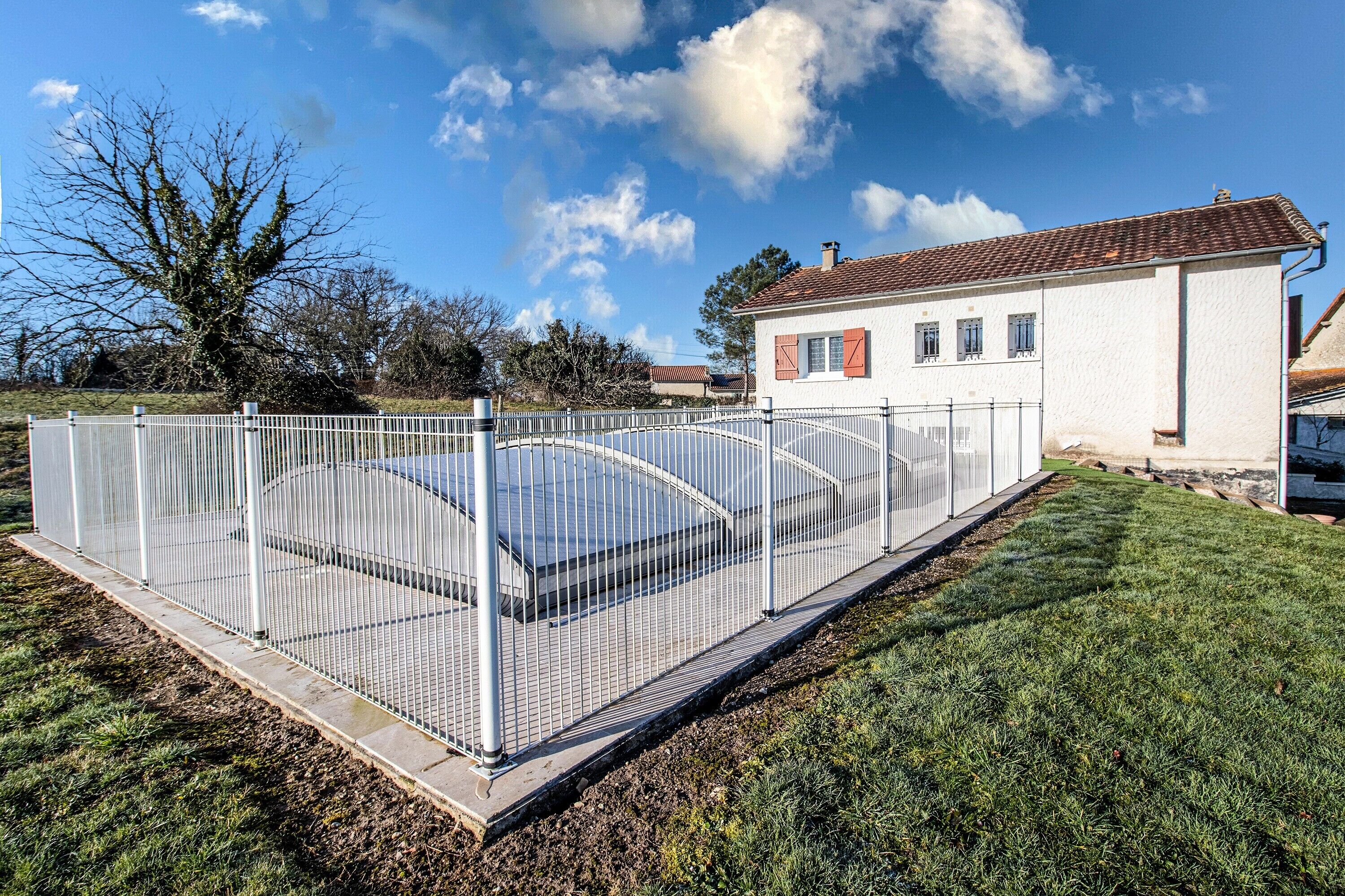 Ein eingezäunter Außenbereich mit Pool, ein weißes Gebäude mit rotem Dach und einem strahlend blauen Himmel.