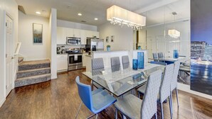 A modern kitchen with stainless steel appliances, a dining area with a glass table and blue chairs, and a cityscape view through a large window.