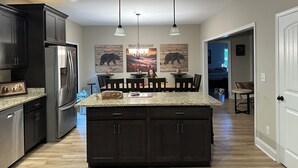 Een moderne keuken met donkere kastjes, roestvrijstalen apparatuur en een centraal keukenblok. Boven het keukenblok hangen hanglampen en er is kunst aan de wanden.