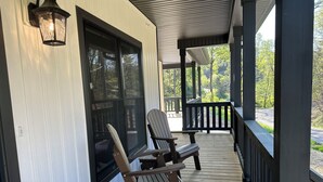 Een houten terras met twee Adirondack-stoelen, een glazen deur en uitzicht op een weg en bomen.