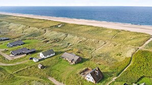 Vista aérea de uma área costeira com vários edifícios, incluindo casas e uma construção maior, rodeada por campos verdes e uma praia.