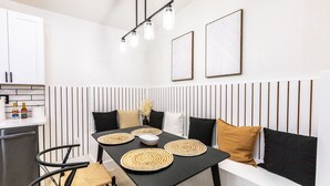 Uma cozinha moderna com área de refeições, apresentando uma mesa preta com jogos americanos trançados, um armário branco e uma luminária pendente com moldura preta.