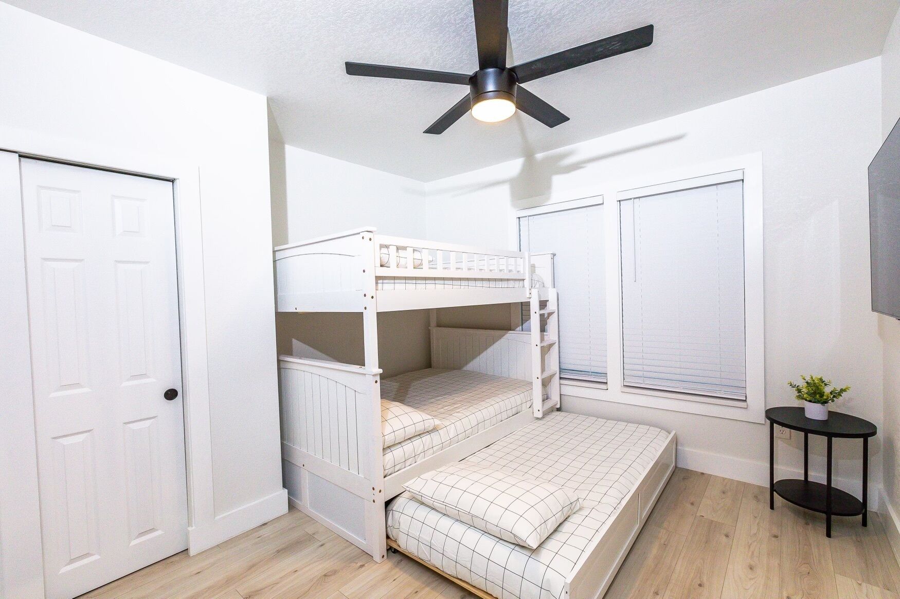 Une chambre avec un lit superposé, un ventilateur de plafond, une porte, une fenêtre avec des stores et une petite table sur laquelle est posée une plante.