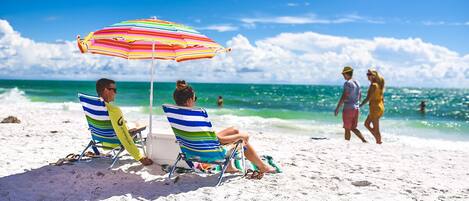 Two people relaxing on beach chairs under a striped umbrella on a sandy beach with the ocean in the background.
