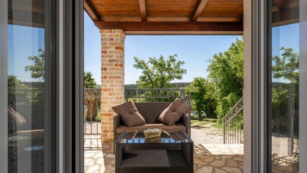 Un canapé en osier agrémenté de coussins, placé sur une terrasse en pierre sous une pergola en bois, avec vue sur des arbres et une clôture en arrière-plan.
