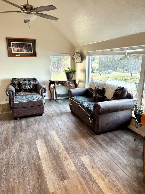 A living room with two sofas, a ceiling fan, a table with a plant, and a window with a view of the outdoors. - Paradise Suites (Homer)