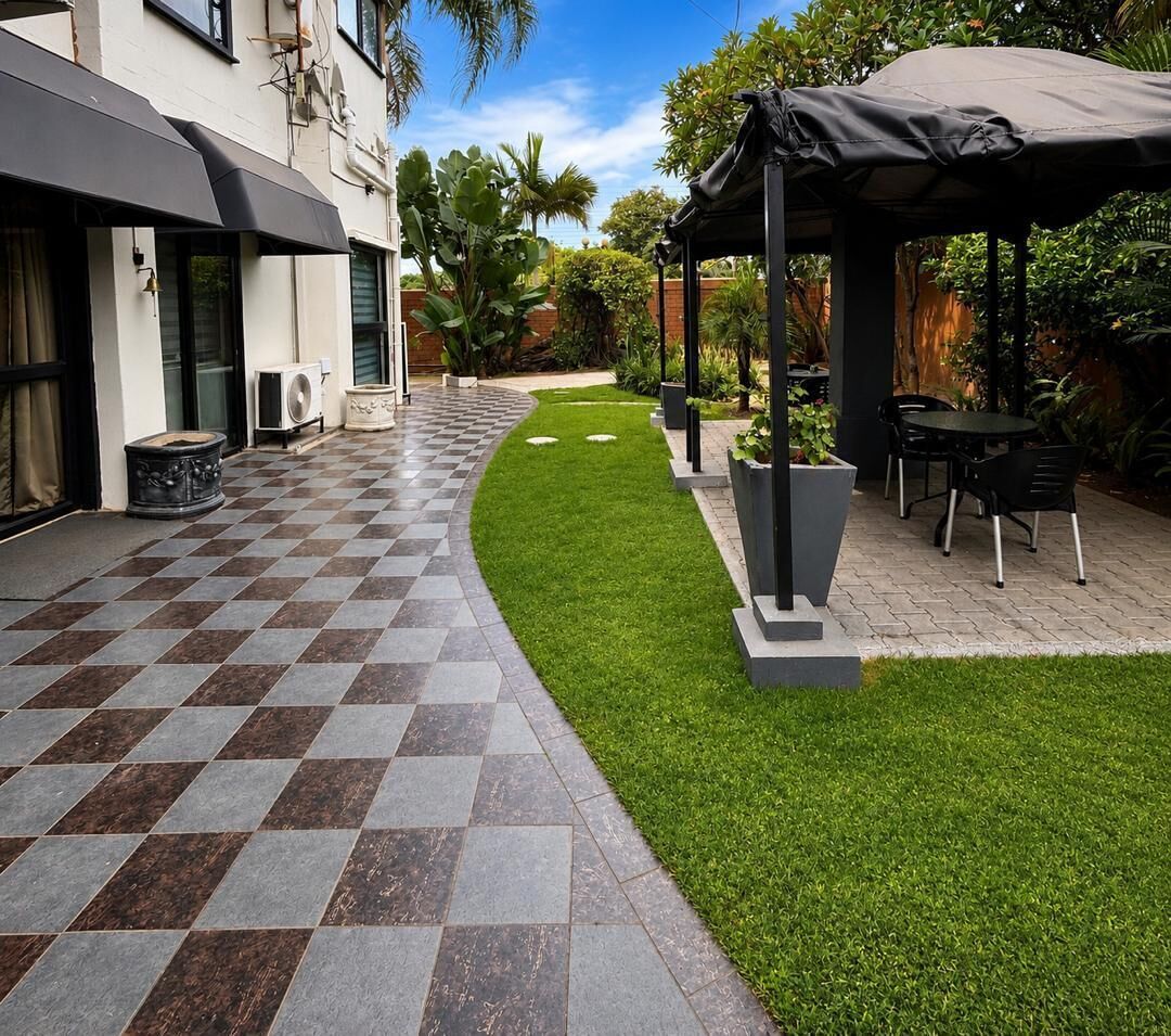 Un patio con piso de baldosas a cuadros, un área verde, un espacio de estar techado y un edificio con toldo negro.