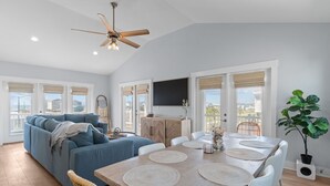 A modern living room with a sofa, dining table, and chairs. There is a ceiling fan, a TV mounted on the wall, and a large window with blinds.