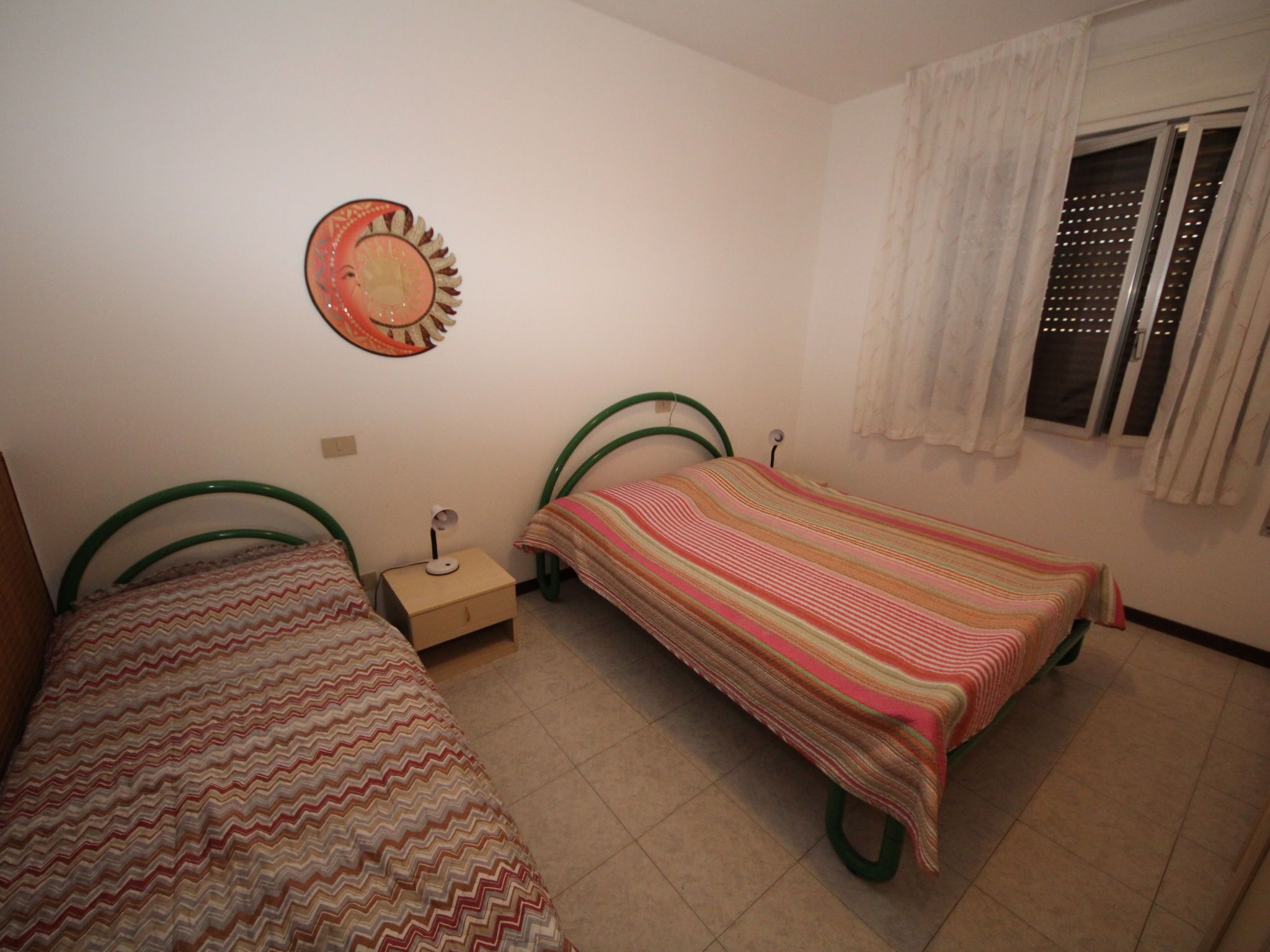 A small, clean room with two single beds, a nightstand, a wall clock, and a window with curtains.