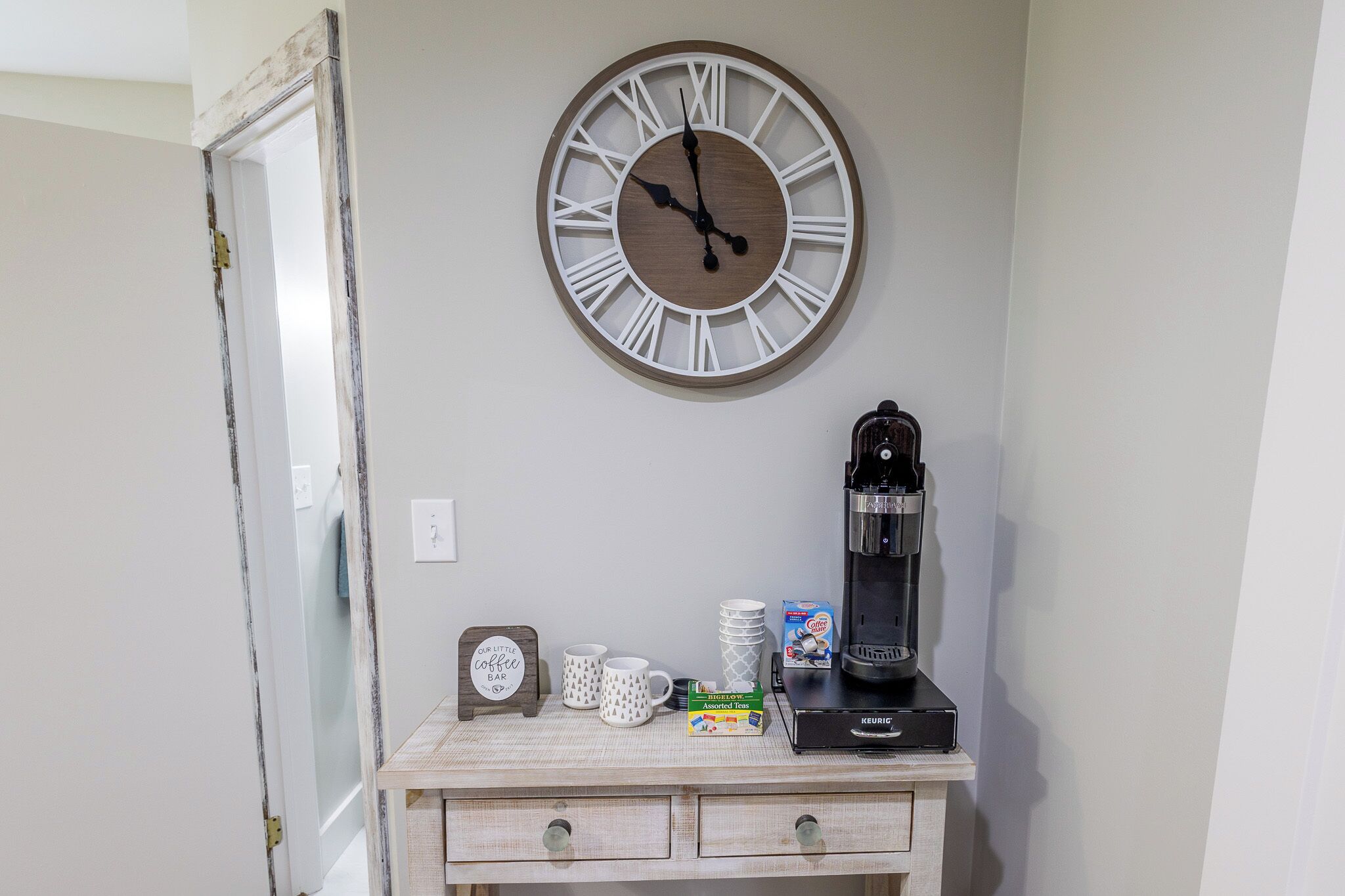 Eine Wanduhr, eine Kaffeemaschine, ein Schild mit der Aufschrift 'Kaffee Bar' und eine Stapel Tassen auf einer Holzkommode.