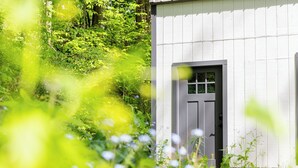 A white wooden cabin with a door and window, surrounded by lush greenery and wildflowers. - Tranquil creek side retreat (Canandaigua)