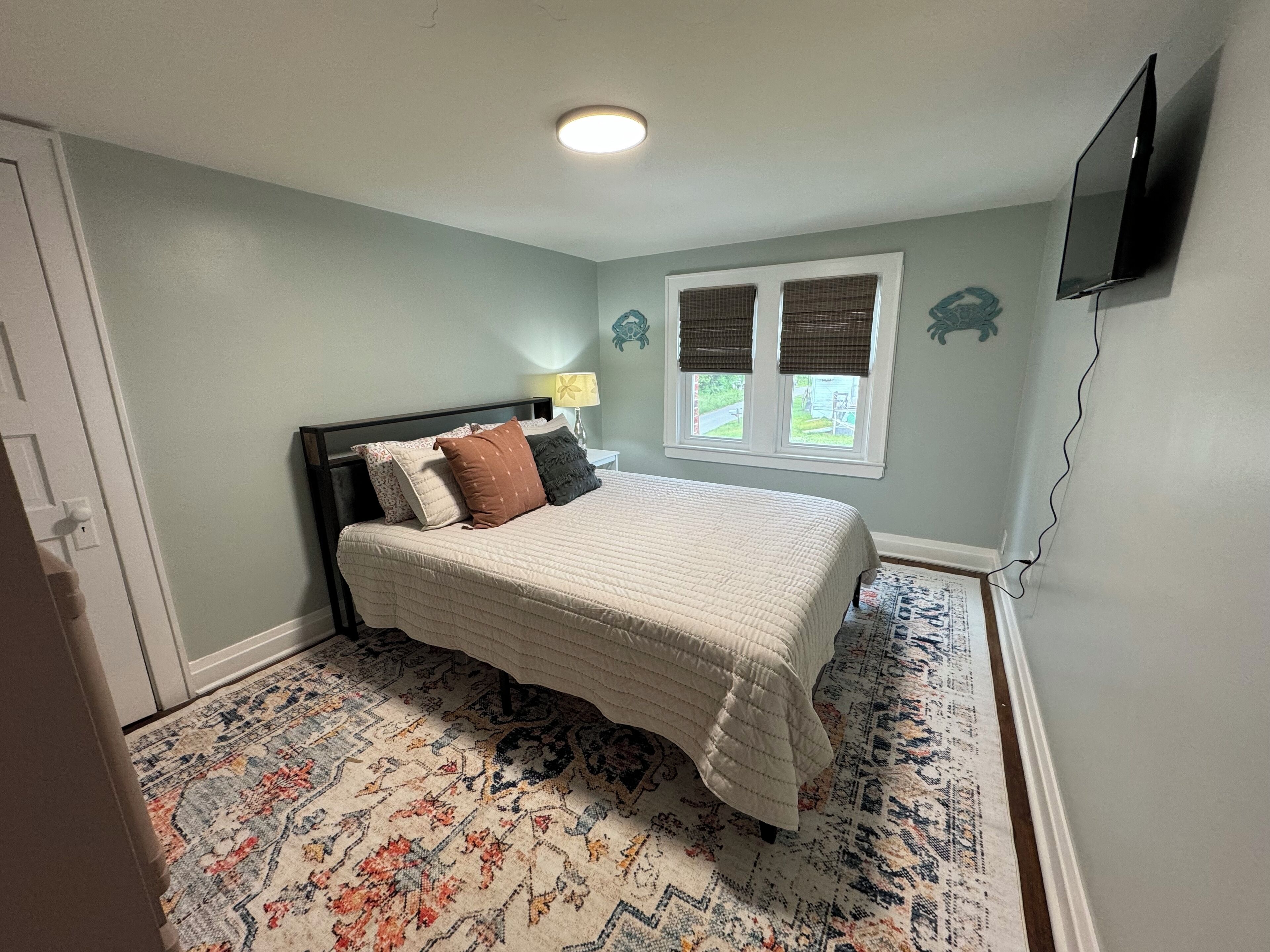 Une chambre à coucher avec un lit, une fenêtre avec des stores, une lampe et un téléviseur fixé au mur.