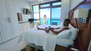 A hotel room with a bed, a TV, a desk, and a view of the ocean.