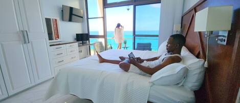 A hotel room with a bed, a TV, a desk, and a view of the ocean.