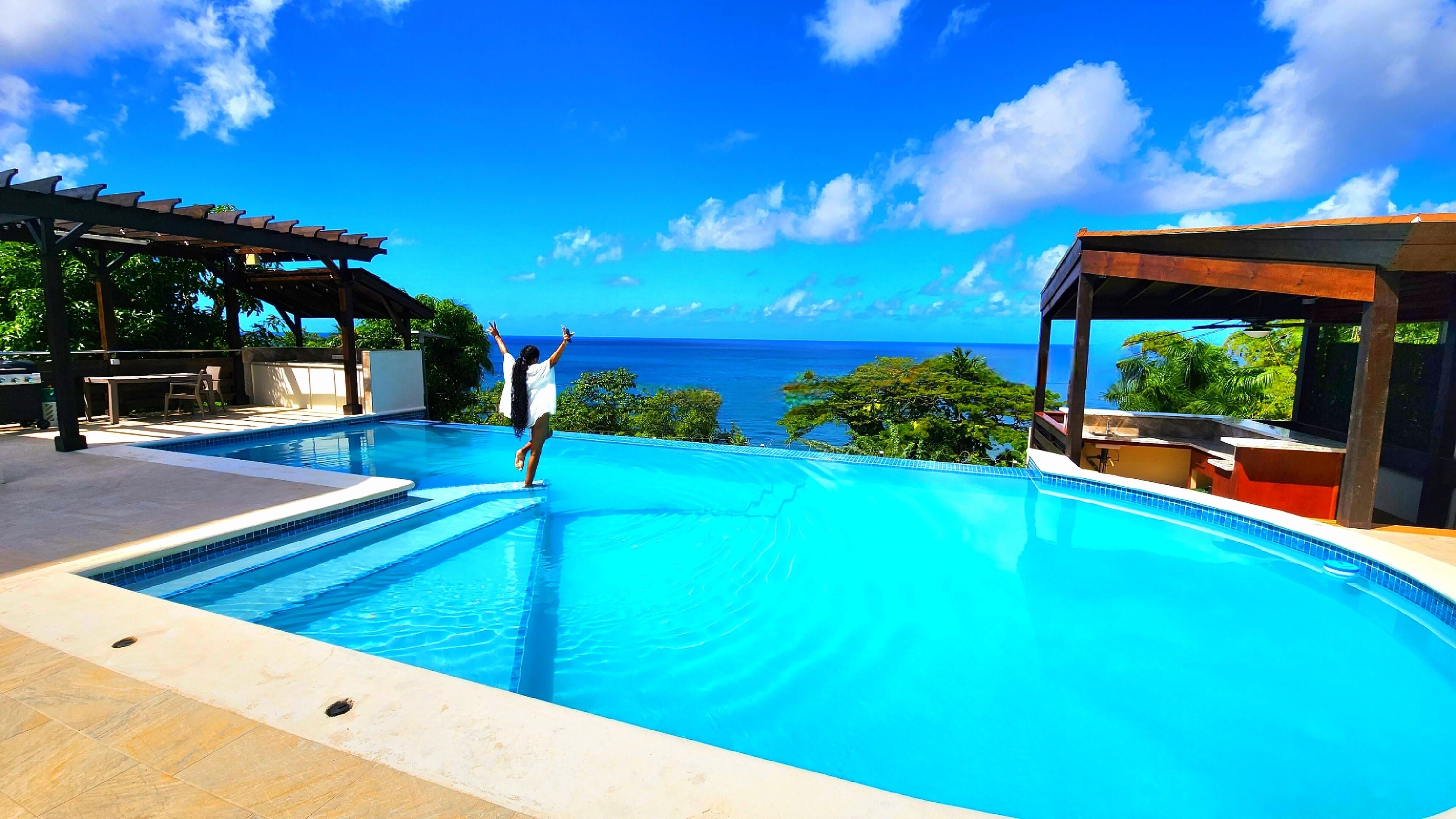 A swimming pool with a view of the ocean, a covered seating area, and a patio with a table and chairs.