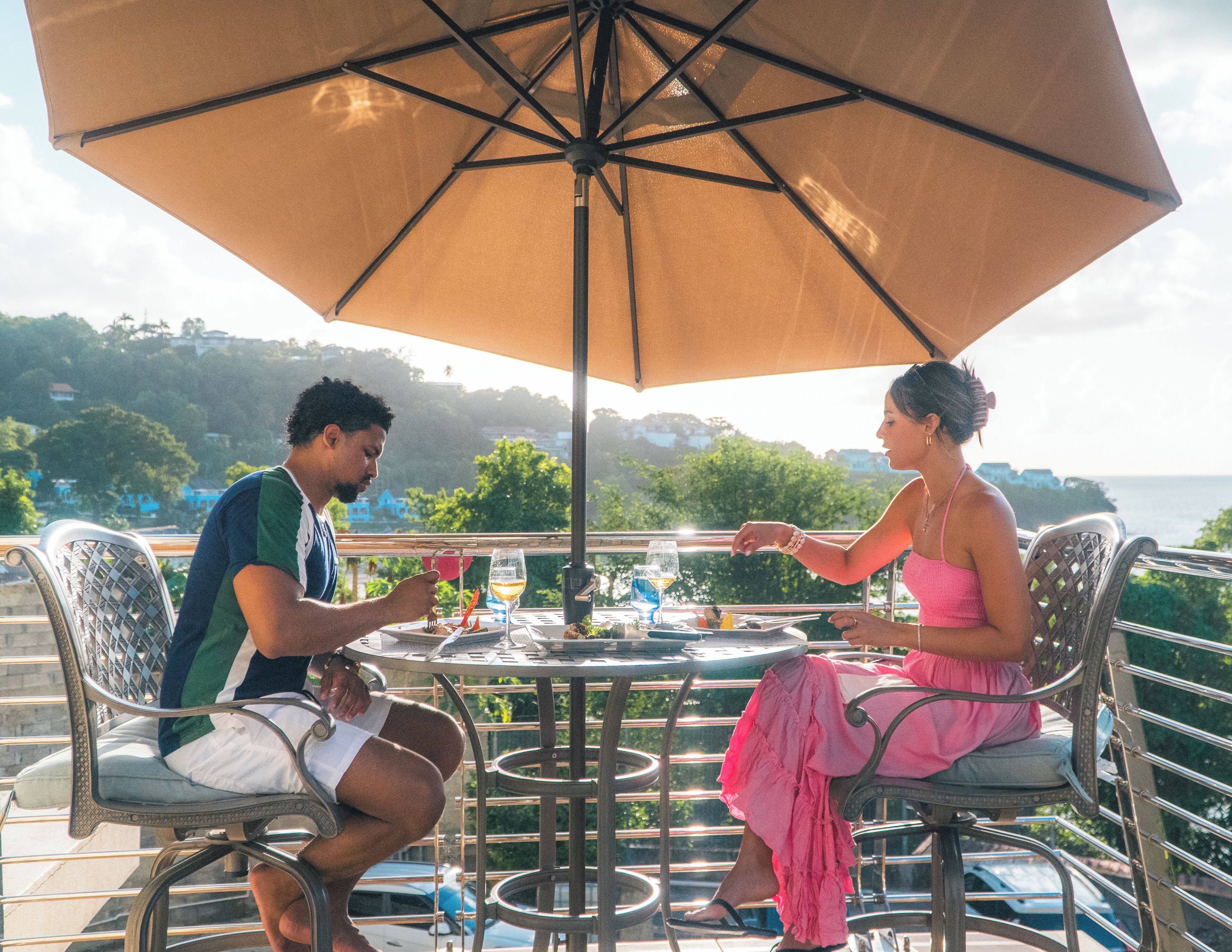 A couple dining at an outdoor table with a large umbrella, overlooking a scenic view with hills and the sea.