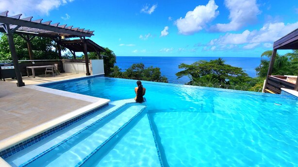 A swimming pool with a view of the ocean, a pergola, and a table with chairs.