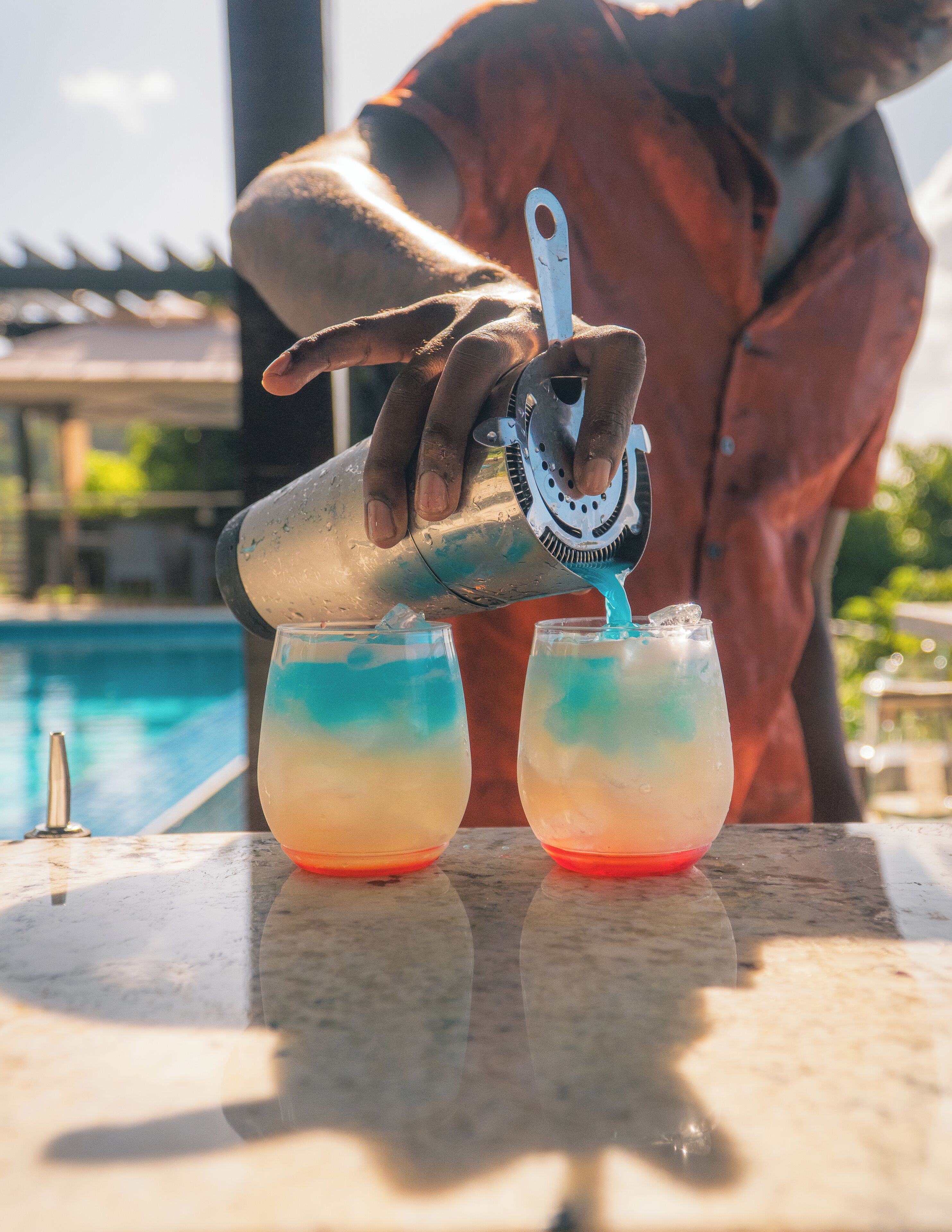 A person pouring a drink into two glasses on a table by a pool.