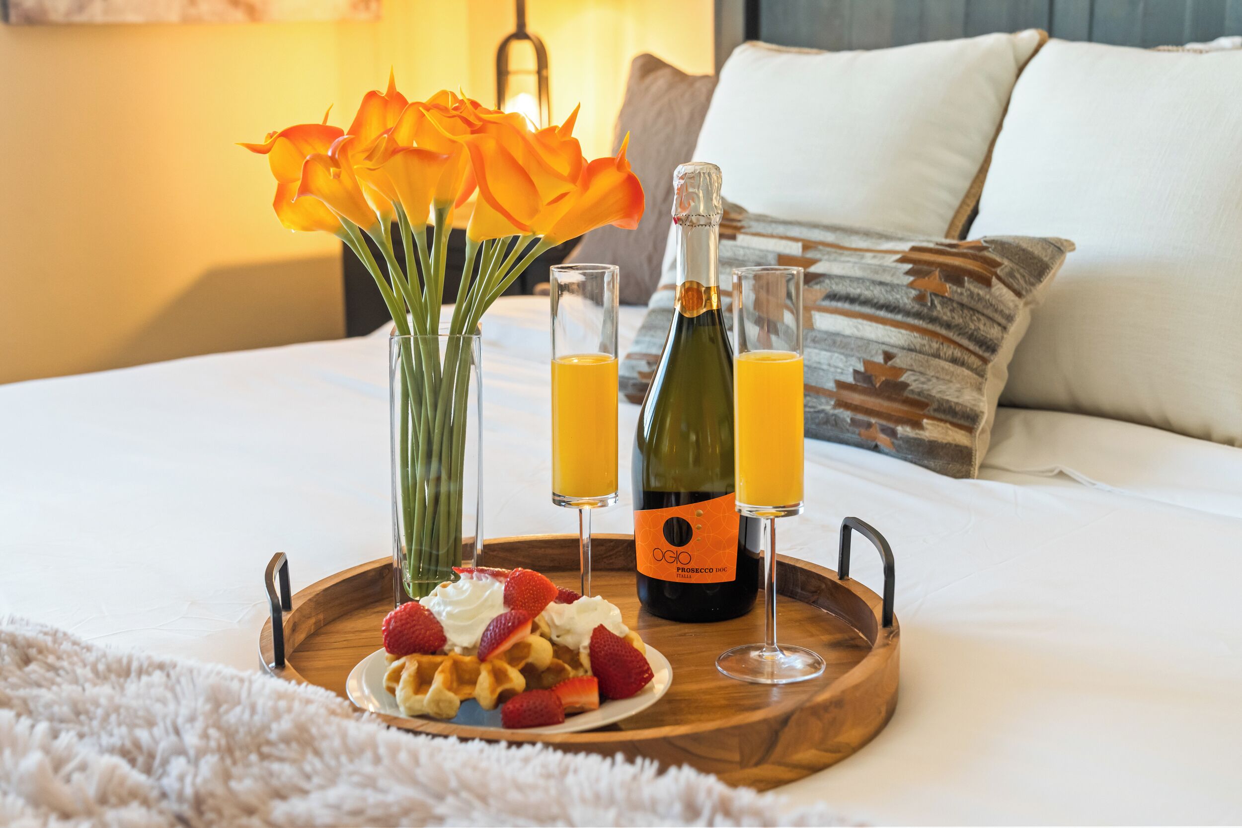 Ein Frühstücksbrett mit Waffeln, Erdbeeren, Schlagsahne und einer Flasche Prosecco auf einem Bett.