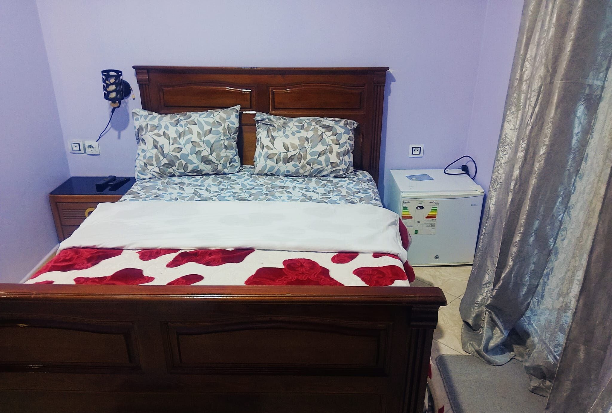 Ein Schlafzimmer mit einem hölzernen Bett, gemusterten Kissen, einer rot-weißen Bettdecke, einem Nachttisch mit Lampe und einem kleinen weißen Gerät am Rand.