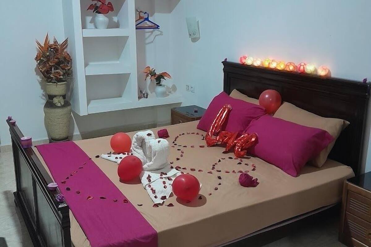 A neatly arranged bedroom with a bed, a bedside table, a nightstand, a white shelving unit, and a potted plant.