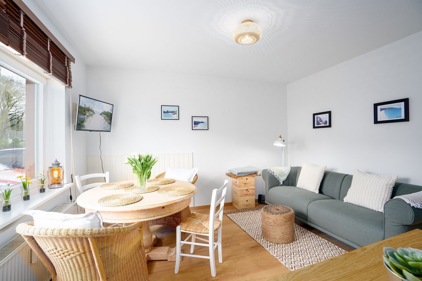 A cozy living room with a round wooden table, white chairs, a grey sofa, and a large window with blinds.
