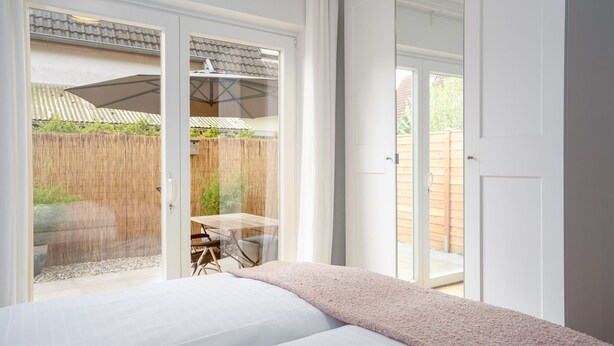 A bedroom with a bed, a sliding glass door leading to an outdoor patio with a table and chairs, and a view of a wooden fence.