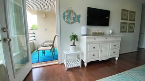 A room with a wooden floor, a white dresser, a small white table with a plant, a fish-shaped sign, and a TV mounted on the wall. - Vacationing is easy in this newly refreshed beach condo! (Virginia Beach)