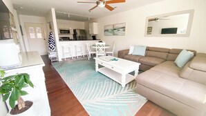 A modern living room with a beige sofa, a white coffee table, and a patterned rug. There is a kitchen area in the background with white cabinets and a dining table. - Vacationing is easy in this newly refreshed beach condo! (Virginia Beach)