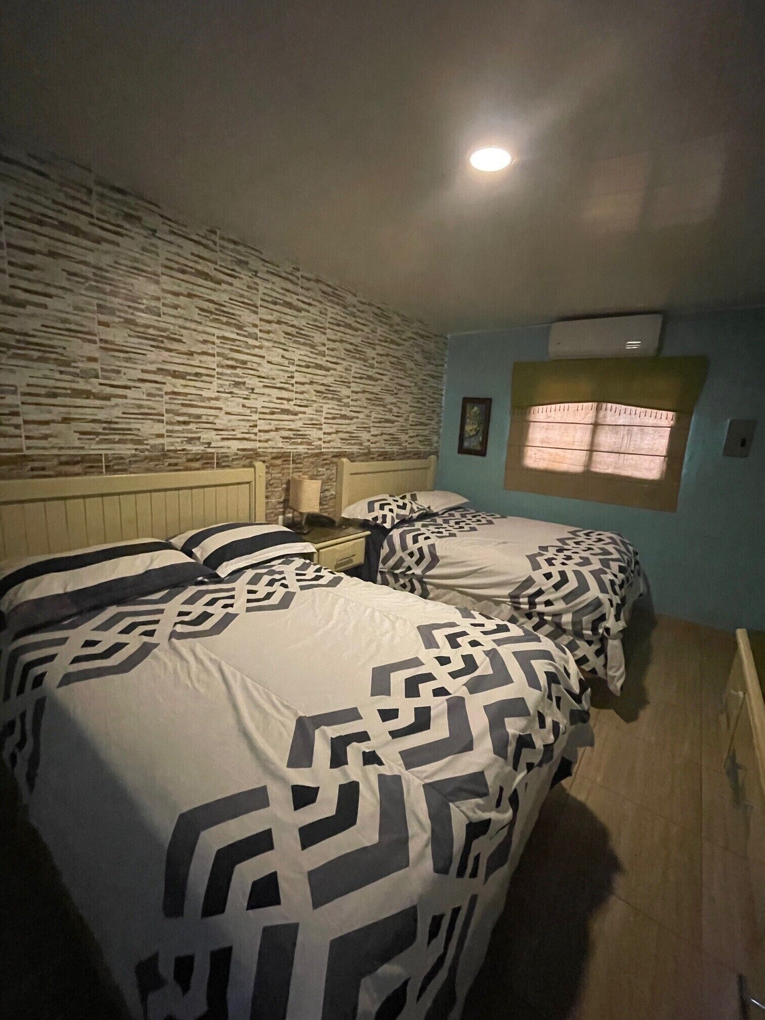 A bedroom with two beds, a stone-patterned wall, a small window with curtains, and a ceiling light.