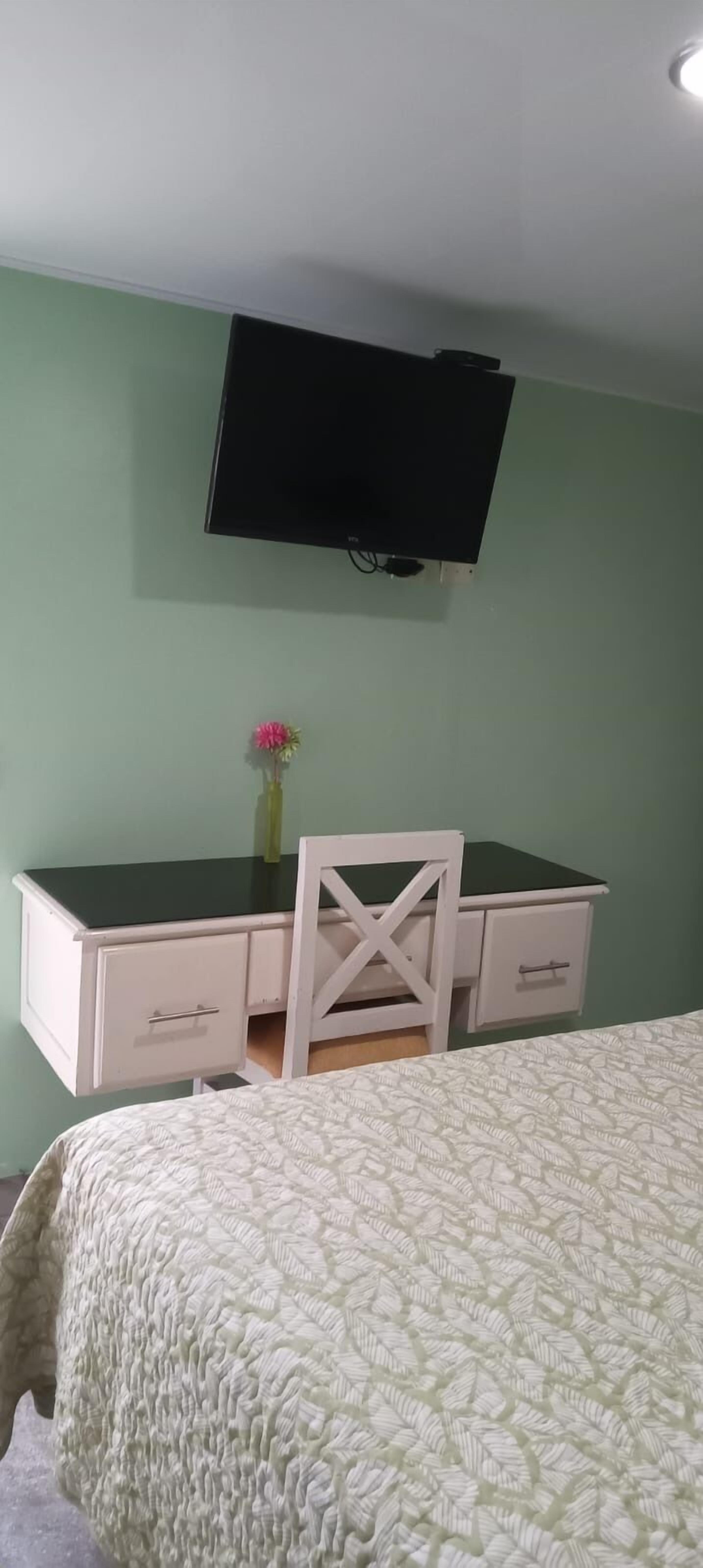 A bedroom with a bed, a desk with drawers, a chair, a television mounted on the wall, and a small vase with flowers on the desk.