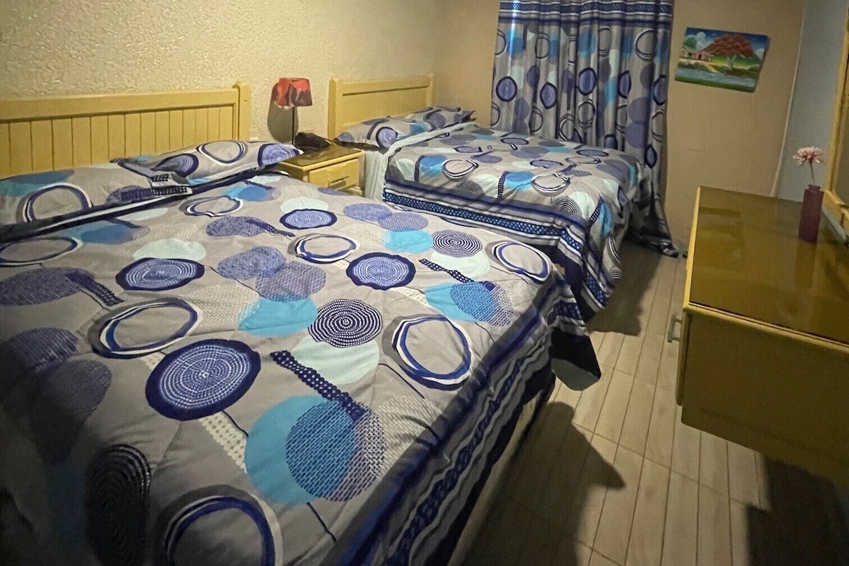 Una habitación con dos camas, una mesita de noche, un equipo de aire acondicionado y una pequeña mesa con un florero.
