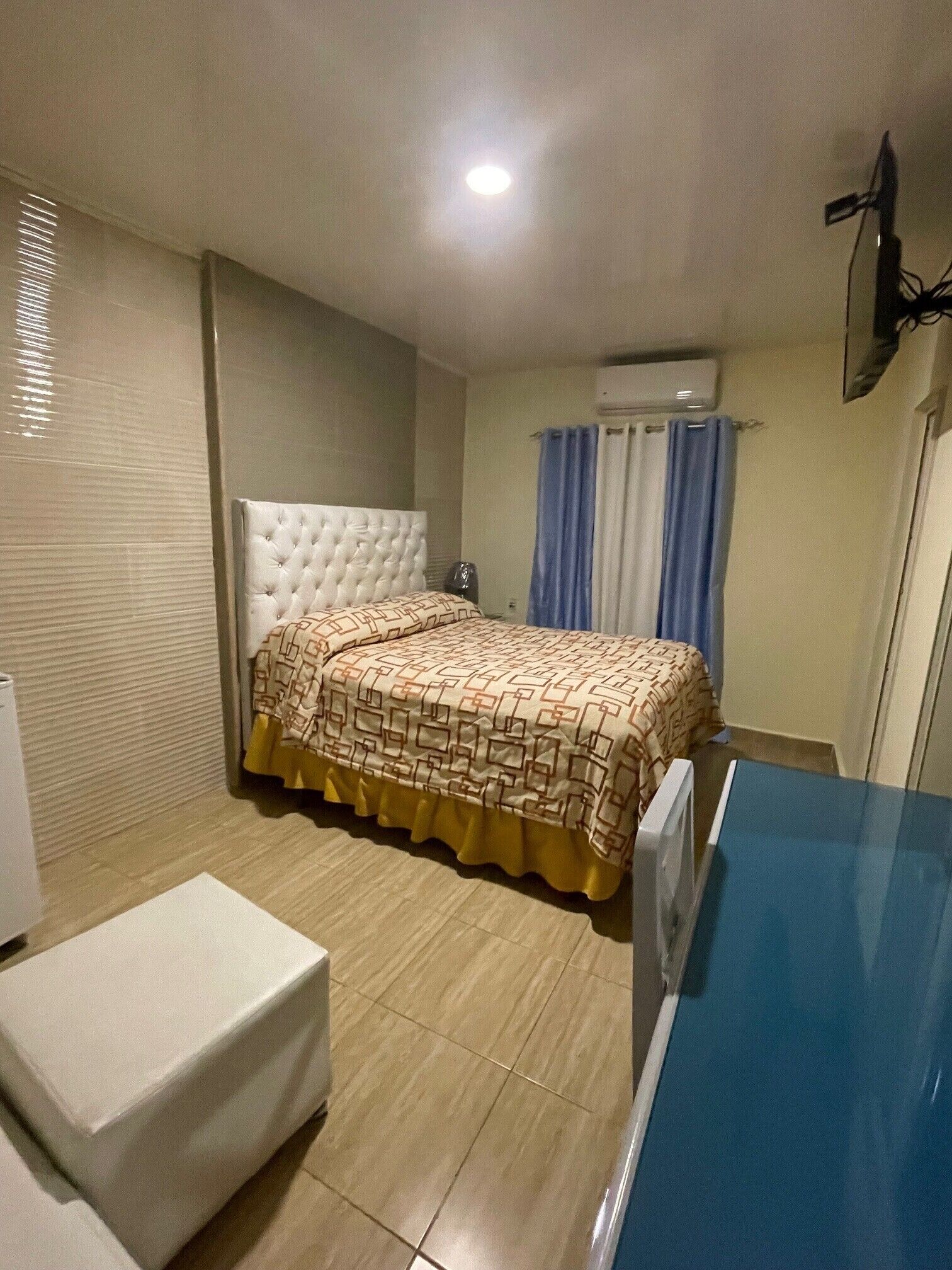 Une chambre à coucher comprenant un lit, une table de chevet, une chaise, un appareil de climatisation et une fenêtre avec des rideaux.
