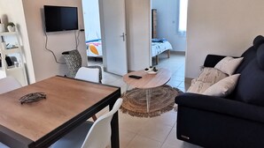 A compact living space with a dining table, a sofa, a TV mounted on the wall, and a bed in the adjacent room. - Between sea and forest 2 near the beaches (Jard-sur-Mer)
