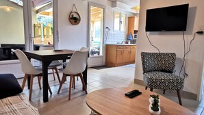 A modern living room with a sofa, a coffee table, and a patterned armchair. There is a television mounted on the wall and a kitchen area visible through an open door. - Between sea and forest 2 near the beaches (Jard-sur-Mer)