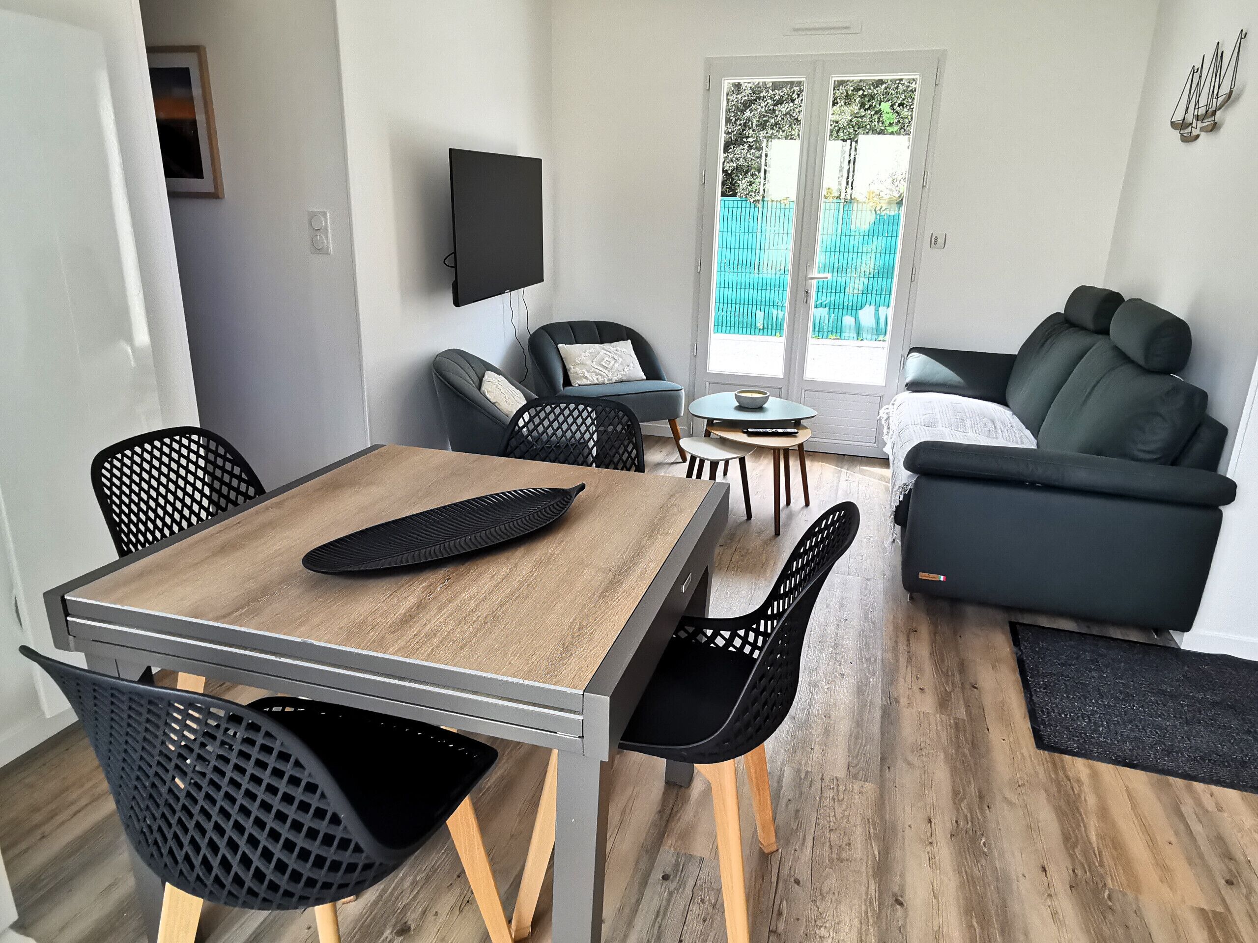 Una sala de estar moderna con comedor, que incluye una mesa de madera, sillas negras, un sofá negro y un televisor montado en la pared.