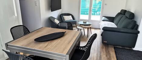 Una sala de estar moderna con comedor, que incluye una mesa de madera, sillas negras, un sofá negro y un televisor montado en la pared.