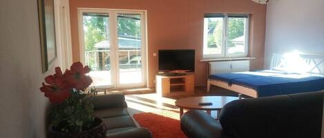 A living room with a sofa, a coffee table, a TV, and a bed. There is a window with a view of trees and a building outside.