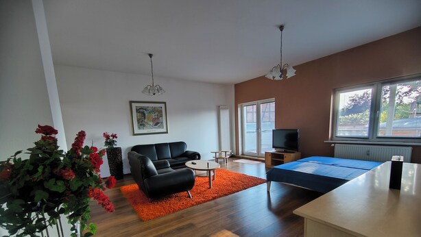 Un salon moderne avec un canapé noir, une petite table ronde, un téléviseur et un lit recouvert d’une couverture bleue.