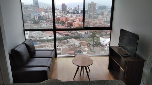 A spacious living room with a large window offering a city view, a comfortable sofa, a small round table, and a TV stand.