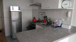 A compact kitchen with a stainless steel refrigerator, a gas stove, and a microwave. There is a clock on the wall and a phone on the counter.