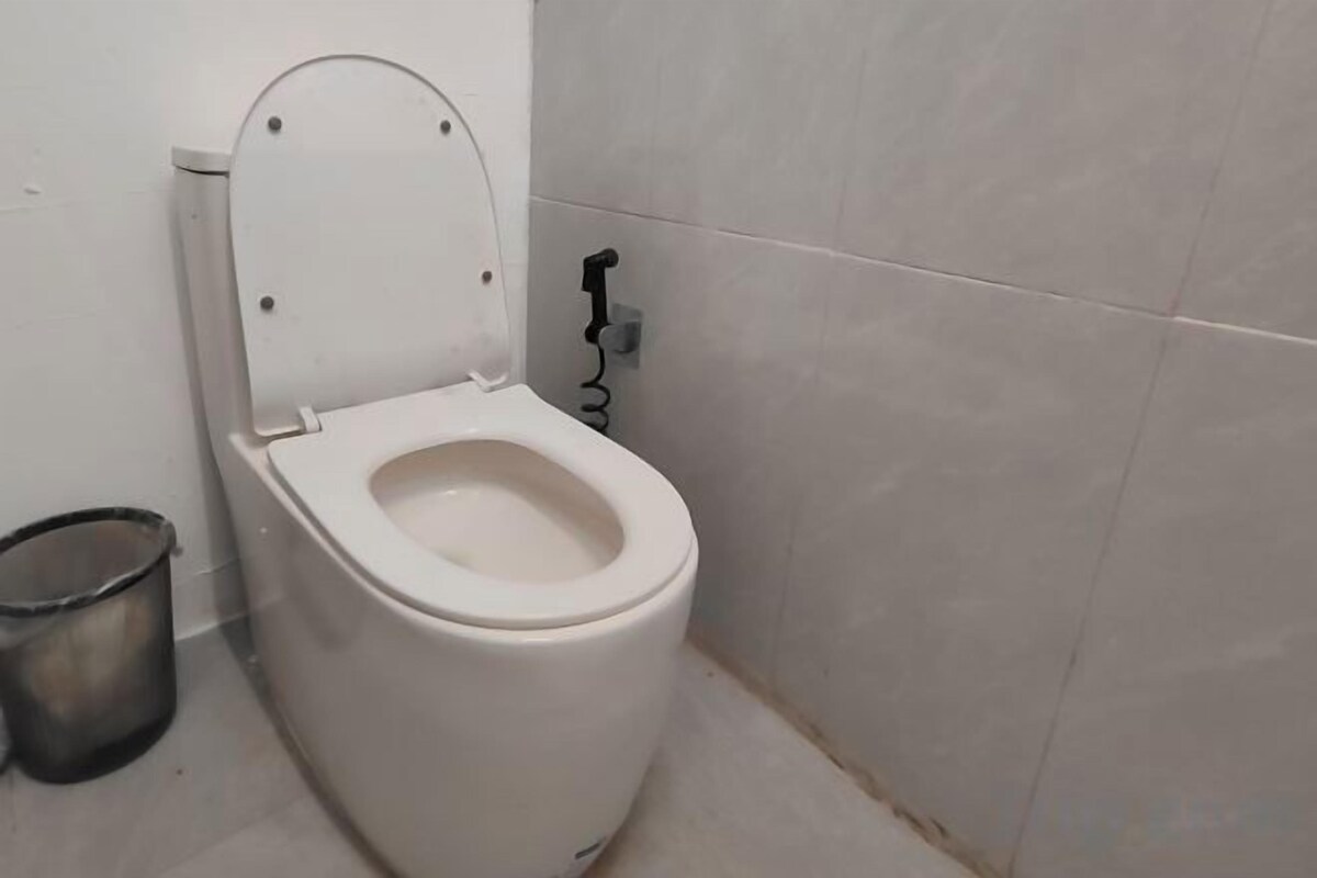 A white toilet with a closed lid, a metal trash bin, and a wall-mounted flush handle.