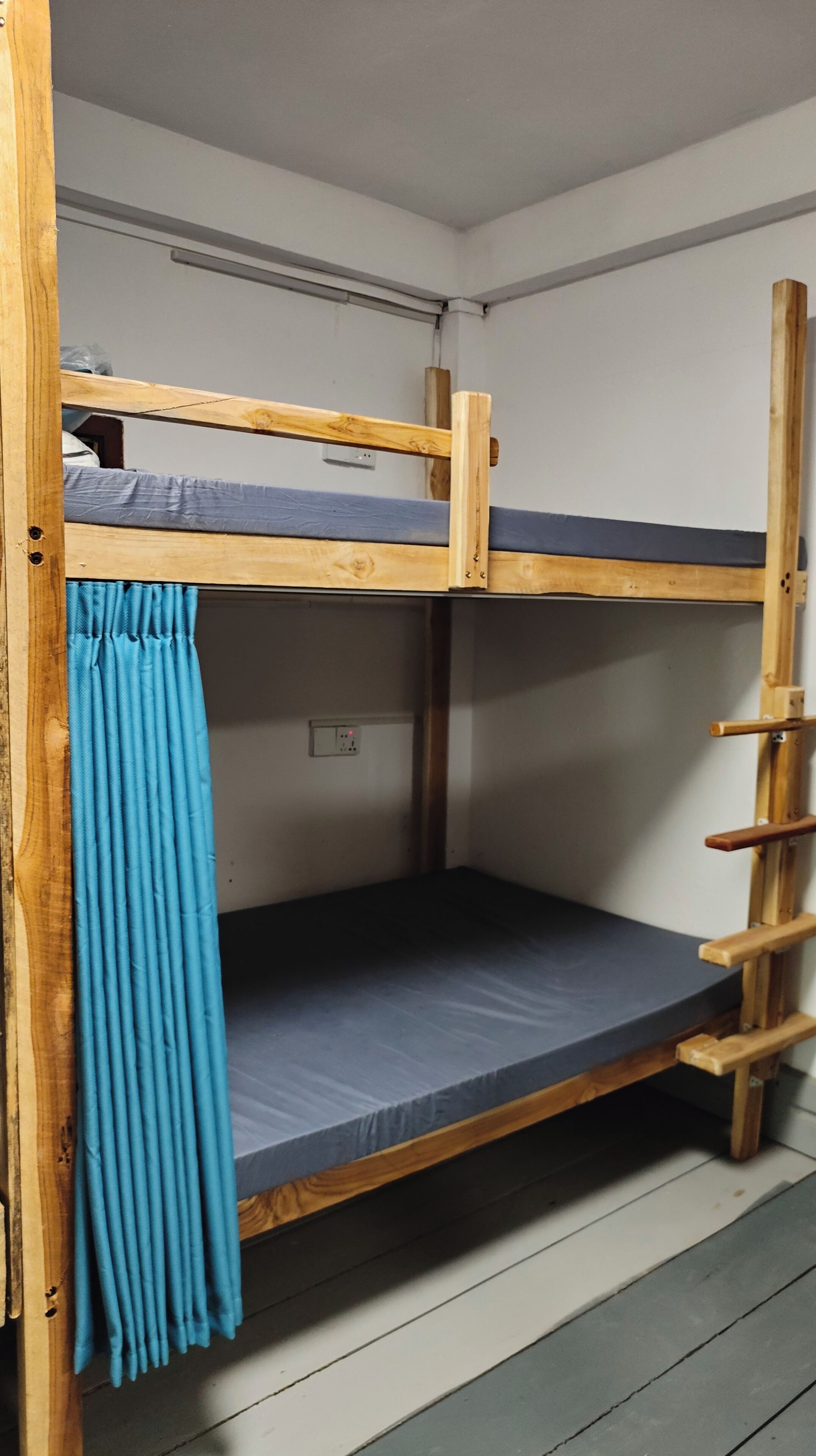 A bunk bed with a blue curtain, a wooden ladder, and a power outlet on the wall.