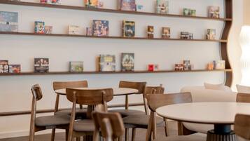 A modern interior with wooden chairs and tables, a bookshelf with various books, and a white wall.