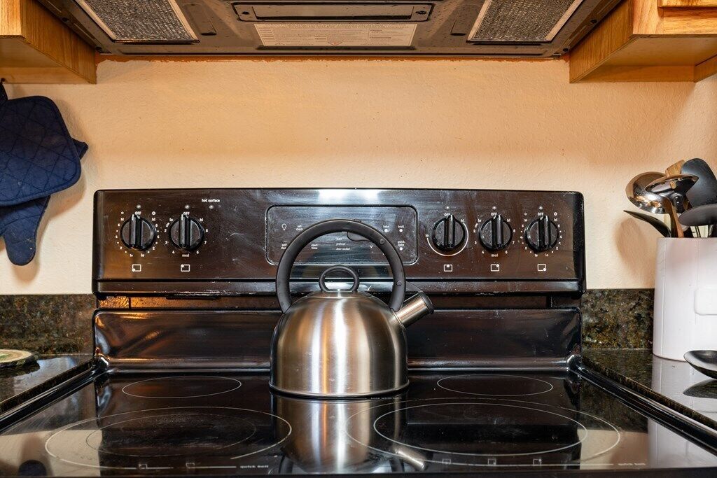 Una cocina con estufa, hervidor y un guante de horno azul en la pared.