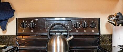 Una cocina con estufa, hervidor y un guante de horno azul en la pared.