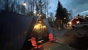 Une petite cabane rustique dotée d’un porche et de deux chaises, entourée d’arbres, avec un bâtiment rouge en arrière-plan.