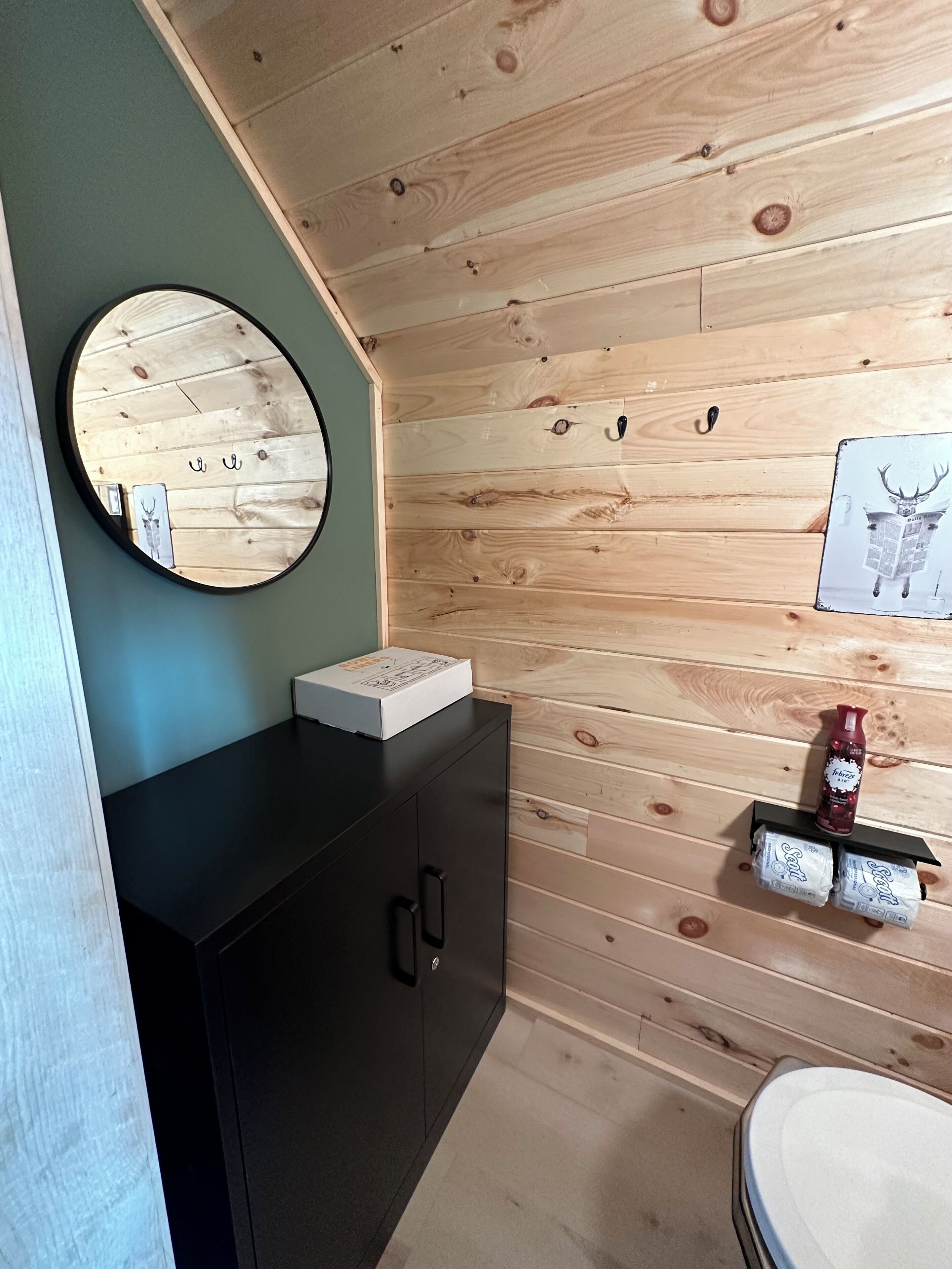 A bathroom with wooden paneling, a round mirror, a black cabinet, and a shelf with toiletries.