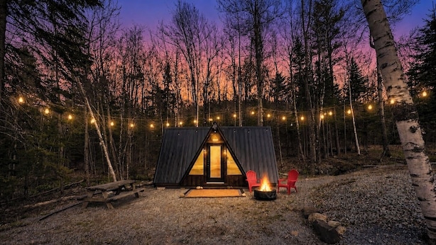 Une cabane rustique avec un toit triangulaire, entourée d’une forêt. On y trouve deux chaises rouges et un foyer à l’extérieur.